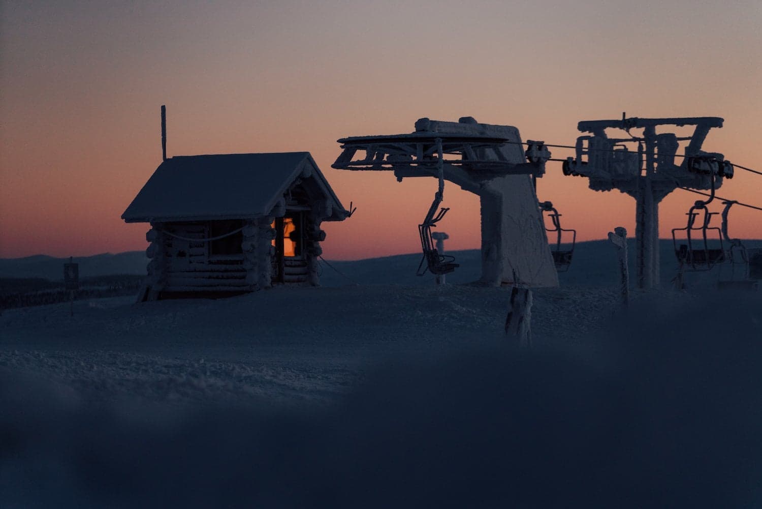 A snow-covered cabin lit from inside at dusk in Finnish Lapland, frost-covered chairlift beside it